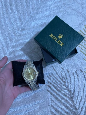  klocka med guld och stenar - Säljer en lyxig  klocka med guldfärgad urtavla och armband dekorerat med glittrande stenar. Klockan har klassisk rund form och levereras i original box. Perfekt accessoar för dig som gillar exklusiv stil.