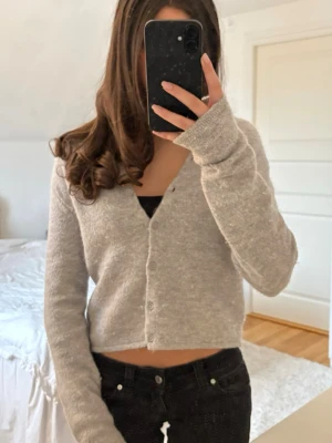 Grå stickad kofta  - Mysig grå kofta i en mjuk, stickad ullblandning. Croppad modell med rund halsringning och knäppning framtill. Perfekt att styla med jeans eller kjol för en avslappnad look.