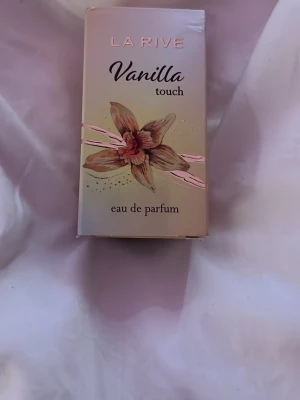 La Rive Vanilla Touch EdP - La Rive Vanilla Touch eau de parfum med en söt och varm vaniljdoft. Flaskan är genomskinlig med guldfärgat lock och etikett, och kommer i en beige kartong med en vaniljblomma på framsidan. Perfekt för dig som älskar mjuka, gourmanddofter. Helt ny och oanvänd. 