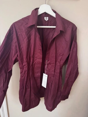 Vinröd skjorta från ARKET - Vinröd skjorta från Arket med klassisk krage och långa ärmar. Skjortan har knäppning framtill och är tillverkad i ett lätt bomullsmaterial. Perfekt för en stilren look och enkel att matcha med olika outfits.