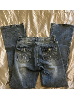 Blå bootcut jeans med dekorerade fickor - Säljer ett par low waist blå bootcut jeans med snygga dekorerade bakfickor och silverfärgade knappar. (Det e några små detaljer på fickorna som har åkt av, men annars bra skick) Dom är stretchiga och bekväma💓