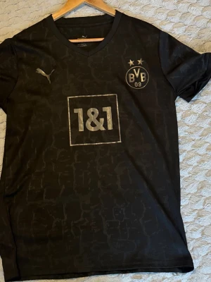Svart Borussia Dortmund tröja Puma - Snygg svart Borussia Dortmund matchtröja från Puma med BVB-logga, två stjärnor och sponsortryck på bröstet. Nummer 11 och namnet Reus på ryggen tillsammans med DORTMUND-tryck. Perfekt för fotbollsfans!