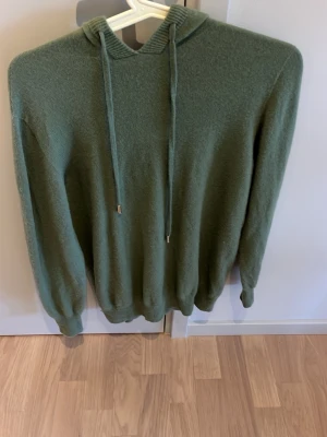 Olivgrön stickad hoodie - Mysig olivgrön hoodie med huva och dragsko. Tillverkad i mjukt, stickat material som ger en avslappnad och skön känsla. Ribbstickade muddar vid ärmslut och nederkant. Perfekt för dig som gillar en enkel och stilren look. Endast använd en gång. Skriv vid funderingar!