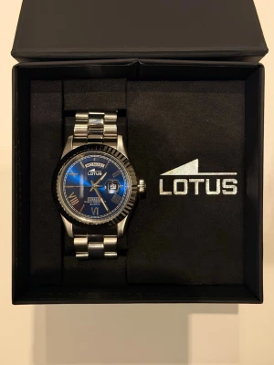 Blå och silver klocka från Lotus - Stilren klocka från Lotus med blå urtavla, silverfärgade romerska siffror och datumvisning. Armbandet är i blankt silverfärgat metall och boetten har en räfflad kant. Perfekt accessoar för dig som gillar klassisk men modern stil.