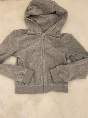 Grå zip-hoodie från Juicy Couture - Snygg grå hoodie från Juicy Couture med dragkedja framtill och två fickor. Klassisk huva och ribbade muddar vid ärmslut och midja. Liten broderad logga på bröstet. Perfekt till jeans eller mjukisbyxor.