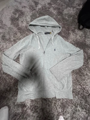 Grå zip hoodie från Polo Ralph Lauren - Snygg gråmelerad hoodie med dragkedja från Polo Ralph Lauren. Tröjan har huva med vita snören, två fickor framtill och den klassiska blå loggan broderad på bröstet. Tillverkad i mjukt bomullsmaterial, perfekt för en avslappnad stil.