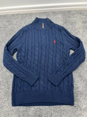 Blå kabelstickad tröja Polo Ralph Lauren - Mörkblå kabelstickad tröja från Polo Ralph Lauren med halv dragkedja och hög krage. Tröjan har långa ärmar, ribbade muddar och den klassiska röda logotypen broderad på bröstet. Perfekt för lager på lager och snygg till jeans.