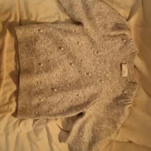 Grå stickad tröja med pärlor Neo Noir - Superfin ljusgrå stickad tröja från Neo Noir med pärldetaljer framtill. Tröjan har rund hals, ribbade muddar och lätt puffade axlar. Perfekt för höst och vinter när du vill ha något både mysigt och snyggt.