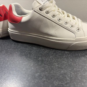 Armani Exchange vita sneakers med röd häl - Snygga vita sneakers från Armani Exchange med röd detalj på hälen och tydlig A|X-logga. Skorna har snörning, rund tå och platt sula. Tillverkade i syntetmaterial med diskret perforerat mönster på sidan. Perfekta för dig som gillar stilrena och trendiga sneakers.