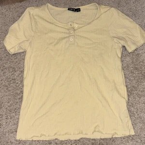 Ribbad beige topp med knappar från LMTD - Säljer en ribbad beige topp från LMTD i storlek XXS. Toppen har korta ärmar, rund halsringning och tre knappar framtill. Den är tillverkad i ett mjukt, stretchigt material och har en enkel, stilren look som passar perfekt till jeans eller kjol.
