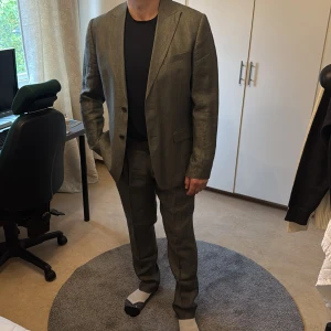 Olivgrön linnékostym med kavaj - Snygg olivgrön kostym i linne med klassisk kavaj och matchande byxor i storlek 52 både i byxor och kavaj. Kavajen har två knappar, spetsiga slag och flera fickor. Materialet är svalt och ger en avslappnad men stilren look. Perfekt för dig som vill ha en modern och fräsch stil. Köpt på MQ market och varan heter nino linen blazer: https://www.mq.se/black-nino-linen-blazer-olive-melange/?gad_source=1&gad_campaignid=22724677855&gbraid=0AAAAACQLh_7OKuUC9MvmQ1GC1mr1kwTEh&scrollToPosition=2304