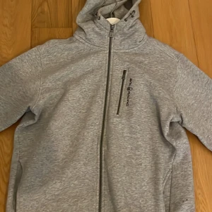 Grå hoodiejacka från Sail Racing XL - Grå hoodiejacka från Sail Racing i storlek XL. Jackan har hel dragkedja framtill, huva med snörning och en bröstficka med dragkedja. Mjuk och sportig känsla, perfekt för chill dagar eller när du vill ha en clean look.