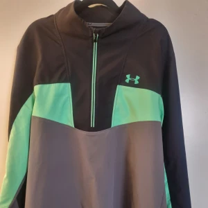 Under Armour vindjacka halfzip - Sportig vindjacka från Under Armour i svart, grått och mintgrönt. Jackan har halv dragkedja framtill, hög krage och logga på bröstet och ryggen.