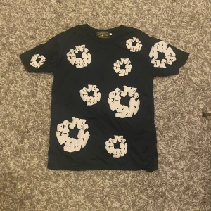 Svart t-shirt med blommigt print Denim Tears - Svart t-shirt från Denim Tears med unikt vitt blommigt print över hela tröjan. Klassisk passform och rund halsringning. Perfekt statement-plagg för dig som vill sticka ut med cool design och streetwear-vibe.
