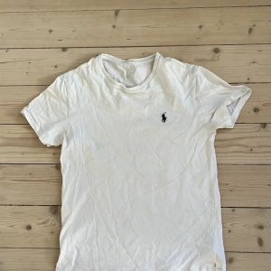 Vit t-shirt från Polo Ralph Lauren - Vit t-shirt från Polo Ralph Lauren med klassisk rund hals och broderad marinblå logga på bröstet. Tillverkad i mjuk bomull och har en enkel, stilren design som passar perfekt till jeans eller shorts.