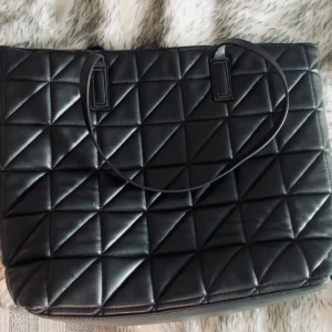 Svart quiltad axelväska i skinnimitation - Stilren svart axelväska med  mönster och långa smala axelband. Väskan är stor, rymlig och har dragkedja upptill samt innerfack med guldfärgad dragkedja. Tillverkad i skinnimitation och passar perfekt för dig som vill ha en trendig och praktisk väska. 