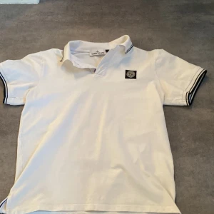 Vit Stone Island piké t-shirt - Vit piké t-shirt från Stone Island med klassisk krage och korta ärmar. T-shirten har svarta detaljer på ärmsluten och kragen samt den ikoniska Stone Island-loggan på bröstet. Tillverkad i mjuk bomull för en skön känsla och snygg passform.