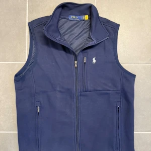 Marinblå väst från Polo Ralph Lauren - Marinblå väst från Polo Ralph Lauren med hög krage, meshfoder och dragkedja framtill. Västen har tre fickor med dragkedja och en vit broderad logga på bröstet. Perfekt för lager-på-lager och sportig stil.