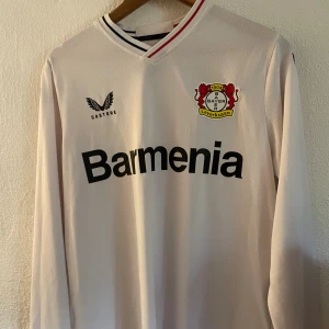 Vit Bayer Leverkusen långärmad tröja - Säljer en vit långärmad Bayer Leverkusen fotbollströja från Castore. Tröjan har klubbmärke på bröstet, svart Barmenia-tryck och snygga detaljer i kragen med rött och svart. Tillverkad i lätt och ventilerande material, perfekt för match eller träning.