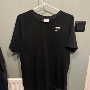 Svart Gymshark t-shirt i polyester - Svart t-shirt från Gymshark i storlek M, perfekt för träning. T-shirten har korta ärmar, rund hals och en stilren logga på bröstet. Materialet är lätt och andas, vilket gör den grym för gymmet eller sport. Snygg och enkel design som funkar till alla träningspass.