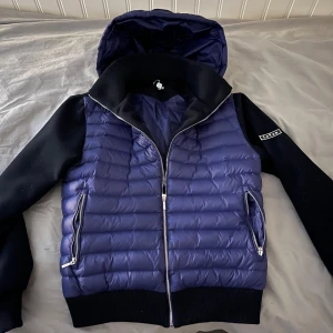 Blå Cardigan Jacka från LeVon - Snygg cardigan jacka från LeVon med blå, quiltad framsida och huva samt svarta ärmar och rygg. Jackan har dragkedja framtill och två dragkedjeförsedda fickor. Perfekt för kyligare dagar. Storlek M