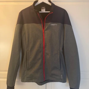 Grå vindjacka från Craft med röd dragkedja - Säljer en grå och mörkblå vindjacka från Craft med snygg röd dragkedja. Jackan har hög krage, två fickor framtill och lång ärm. Perfekt för dig som gillar sportig stil och vill ha något lätt och funktionellt. Storlek M