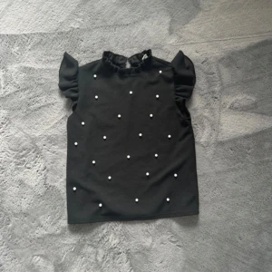 Svart blus med pärlor från SHEIN - Supersöt svart blus från SHEIN med volangkrage och korta puffärmar. Framsidan är dekorerad med små vita pärlor som ger en extra touch. Blusen har en liten knapp i nacken för enkel påklädning. Perfekt för dig som gillar detaljer och stil.