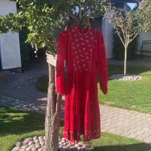 Röd långklänning med paisleymönster - Röd långklänning med paisleymönstrat liv och nederkant, höga axlar och lång ärm. Klänningen har ståkrage, dekorativa gulddetaljer längs livet och dragkedja framtill. Materialet känns som bomull och modellen är luftig och flowy.