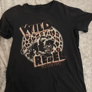 Svart t-shirt Wild Rebel Gina Tricot XS - Svart t-shirt från Gina Tricot i storlek XS med tryck av en panter, texten 'Wild Rebel Tour of 1979' och leopardmönstrad bakgrund. Klassisk rund hals och korta ärmar. Skön bomullskvalitet och coolt statement-tryck framtill.