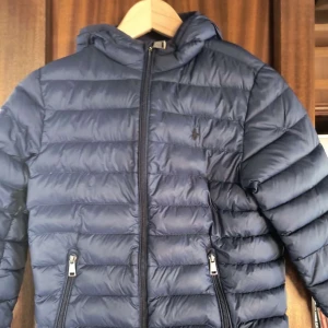 Mörkblå dunjacka Polo Ralph Lauren - Snygg mörkblå dunjacka från Polo Ralph Lauren med huva och dragkedja framtill. Jackan har två fickor med dragkedja och klassisk quiltad design. Perfekt för kyliga dagar och riktigt stilren look.