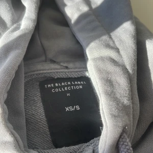 Grå hoodie från The Black Label Collection - Snygg grå hoodie från The Black Label Collection i storlek XS/S. Tröjan har huva med snörning, magficka och ribbade muddar. Perfekt för chill dagar och enkel att matcha med jeans eller joggers.