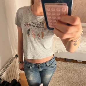 Grå croppad t-shirt med fjädertryck - Säljer en ljusgrå croppad t-shirt med korta ärmar och tryck av en pil med hängande fjädrar i rosa, brunt och svart. Toppen har en avslappnad passform och är tillverkad i mjukt bomullsmaterial. Perfekt för en chill och trendig look.