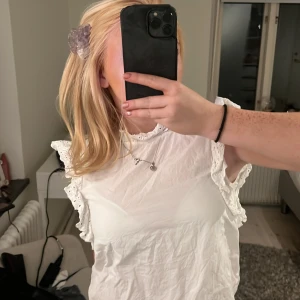 Vit blus med volangdetaljer - Säljer en söt vit blus med volangkantade ärmhål och hög hals. Blusen har broderade spetsdetaljer längs volangerna och är ärmlös, perfekt för en fräsch och feminin look. Passar till både jeans och kjol., blir såklart snyggare om den stryks! ❤️