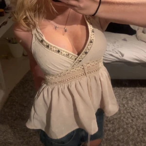 Beige omlottopp med spetsdetaljer - Supersöt beige omlottopp med djup v-ringning och spetsdetaljer längs urringningen och midjan. Toppen har knyt i midjan och ett lätt, luftigt tyg som faller fint. Perfekt för dig som gillar romantisk och feminin stil.