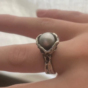 Silvrig ring med pärla - Unik och chunky ring i silverfärgad metall med en stor vit pärla i centrum. Ringen har en organisk, skulptural form med grov yta som ger den en cool och edgy vibe. Perfekt statement piece för dig som gillar att sticka ut. Pris går att diskutera💗💗