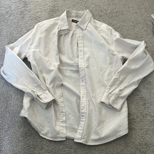 Vit randig skjorta från Dressmann - Klassisk vit skjorta från Dressmann med tunna grå ränder. Skjortan har lång ärm, knappar framtill och krage. Perfekt för en clean och stilren look. Tillverkad i mjuk bomull som känns skön mot huden.