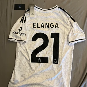 Nottingham Forest Elanga tröja - Snygg vit kortärmad Nottingham Forest fotbollströja från Adidas med svart tryck, Elanga #21 på ryggen och klubbmärke på bröstet. Tröjan har svarta detaljer, tre ränder på axlarna och ett unikt mönster över hela materialet. Perfekt för dig som gillar Premier League.