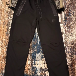 Svarta Nike mjukisbyxor med dragkedjor - Svarta mjukisbyxor från Nike med smal passform, dragsko i midjan och två fickor med dragkedja framtill. Byxorna har diskret Nike-logga på ena benet och är tillverkade i mjukt syntetmaterial som passar perfekt för träning eller chill. Storlek L känns som m