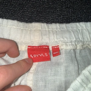 Vita linnebyxor från RYVLS - Säljer ett par vita linnebyxor från RYVLS med elastisk midja och raka ben. Byxorna har en avslappnad passform och är tillverkade i svalt linnematerial, perfekta för varmare dagar. Klassisk look med fickor i sidorna och enkel design.