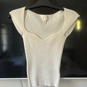 Vit ribbad topp från H&M XS - Snygg vit ribbad topp från H&M i storlek XS. Toppen har en figurnära passform, breda axelband och markerad byst med coola sömdetaljer. Tillverkad i stretchigt material som sitter skönt och framhäver siluetten. Perfekt att styla med jeans eller kjol. Använd 1 gång
