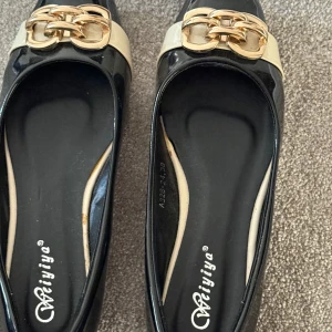 Svarta ballerinaskor med gulddetalj Weiyiya - Snygga svarta ballerinaskor från Weiyiya med lackad finish och breda beige band framtill. Skorna har en fyrkantig tå och är dekorerade med stora guldfärgade kedjedetaljer som ger en lyxig känsla. Perfekta för dig som vill sticka ut med stilrena detaljer.