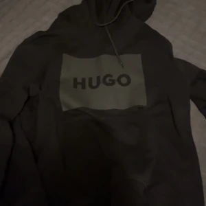 Svart hoodie från HUGO med logga - Svart hoodie från HUGO med stor logga framtill i grått. Klassisk modell med huva och dragsko, samt långa ärmar. Tillverkad i mjuk bomullsmix som är skön att bära. Perfekt för en chill och stilren look.