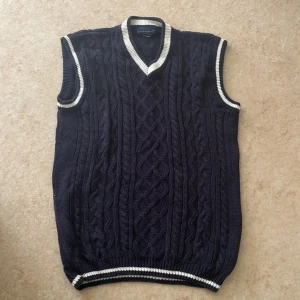 Mörkblå stickad väst från Cotton Republic - Snygg mörkblå stickad väst med vita ribbade detaljer vid v-ringad hals, ärmslut och nederkant. Klassisk kabelstickad design som ger en preppy vibe. Perfekt att styla över en skjorta för en cool look.