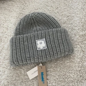 Grå stickad mössa från Marimekko - Unused Marimekko grey “Tilaisuus Solid” beanie. 100% wool. The wool in this product is certified and responsibly produced. Original price: €130.      —- Snygg grå ribbstickad mössa från Marimekko i 100% ull. Mössan har en bred uppvikt kant och framtill sitter en broderad vit blomma på en fyrkantig patch. Perfekt accessoar för kyliga dagar och en stilren detalj till din outfit. 