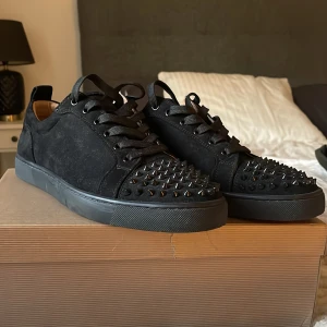 Christian Louboutin svarta sneakers med nitar - Svarta sneakers från Christian Louboutin i mocka med coola svarta nitar på tån och klassisk röd sula. Skorna har snörning och en platt sula, Perfekta för dig som vill sticka ut med exklusiv design och detaljer. 