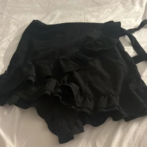 Svart omlottkjol med volanger Bik Bok XS - Supersnygg svart kort omlottkjol från Bik Bok i storlek XS. Kjolen har volanger längs kanten och knytband i midjan. Det är shorts i tjolen. Hör av er vid frågor/intresse💕