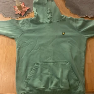 Grön hoodie från Lyle & Scott Junior - Snygg grön hoodie från Lyle & Scott Junior med klassisk gul logga på bröstet. Tröjan har huva, magficka och ribbade muddar. Perfekt för dig som gillar en enkel och stilren look med streetkänsla.