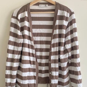 Randig kofta från Cappuccini - Snygg kofta från Cappuccini med breda bruna och vita ränder. Långärmad modell med knappar framtill och ribbade muddar. Perfekt att slänga över en t-shirt för en chill look.