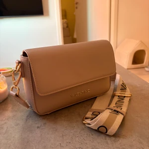 Beige axelväska från Valentino - Snygg beige axelväska från Valentino med guldiga detaljer och bred axelrem med logga. Väskan har flera fack med dragkedja och magnetlås. 