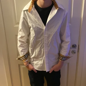 Vit skjorta b - Snygg vit skjorta med klassiska rutiga detaljer på insidan av kragen och ärmsluten. Skjortan har knappar framtill och lång ärm, perfekt för dig som vill ha en clean look med en twist. Tillverkad i bomull för skön känsla. Är storlek L men sitter som M
